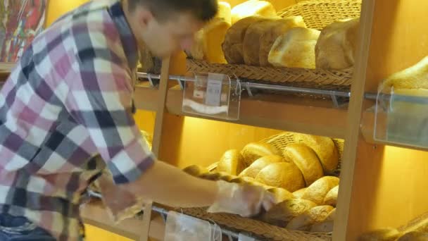 El hombre elige un pan fresco en el supermercado. Un joven toma una barra de pan de la estantería y la huele. Compras en el supermercado — Vídeos de Stock