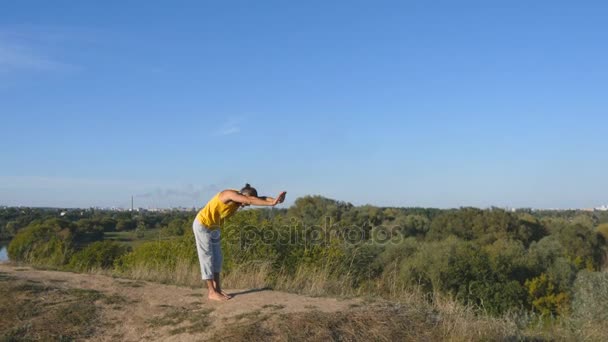Молодой человек, стоящий в позе йоги на природе. Парень практикует йогу и позиционирует себя на улице. Спортсмен делает силовые упражнения на холме. Пейзаж на заднем плане. Здоровый активный образ жизни. Закрыть — стоковое видео