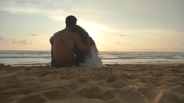 ロマンチックなカップルは、ビーチに座っている美しい夕日とハグを楽しんでいます。女性と男性一緒に座る、海岸の砂の上海や風景を眺めします。スローモーションのクローズ アップ — ストック動画