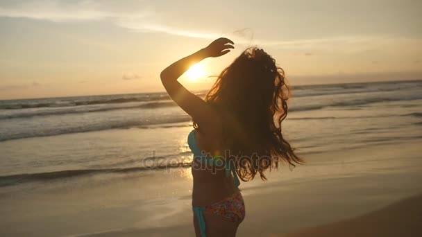 Vacker ung kvinna i bikini står i havet på sunset. Kvinna på stranden njuter av livet under semester. Attraktiva sexig tjej med långt hår poserar på stranden i soluppgången. Slow motion — Stockvideo