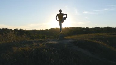 Yoga yapan sporcu silueti dışarıda poz veriyor. Yogi doğada yoga hareketleri ve pozisyonlar üzerinde çalışıyor. Sporcu tek bacağıyla dengede duruyor. Arka plan olarak güzel gökyüzü ve güneş. Sağlıklı aktif yaşam biçimi