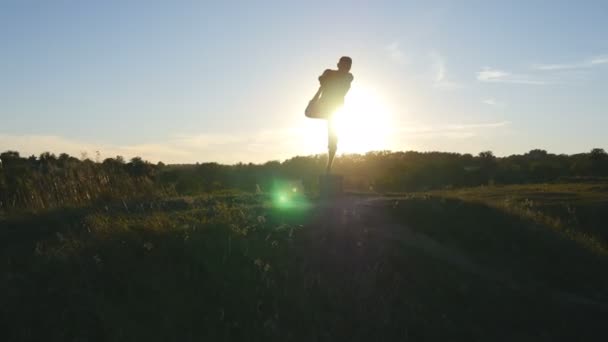 Yogui practicando movimientos de yoga y posiciones en la naturaleza. Silueta de hombre deportivo de pie en la pose de yoga al aire libre. Atleta balanceándose en una pierna. Hermoso cielo y sol como fondo. Vida activa saludable — Vídeo de stock