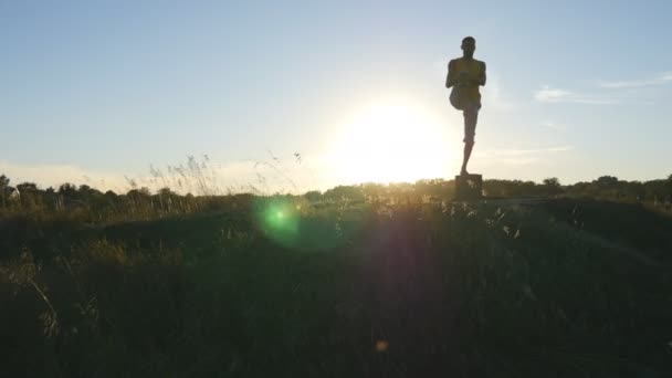 Yogui practicando movimientos de yoga y posiciones en la naturaleza. Silueta de hombre deportivo de pie en la pose de yoga al aire libre. Atleta balanceándose en una pierna. Hermoso cielo y sol como fondo. Vida activa saludable — Vídeos de Stock