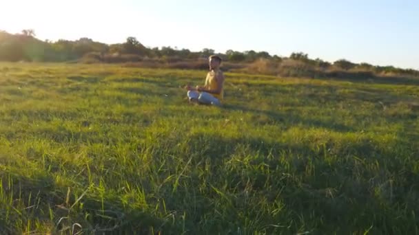 Молодий чоловік, сидячи в йозі, позує на зеленій траві на лузі і медитує. М'язистий хлопець розслабляється в позі лотоса на природі. Відпочинок на свіжому повітрі в сонячний день. Красивий пейзаж на фоні. крупним планом — стокове відео