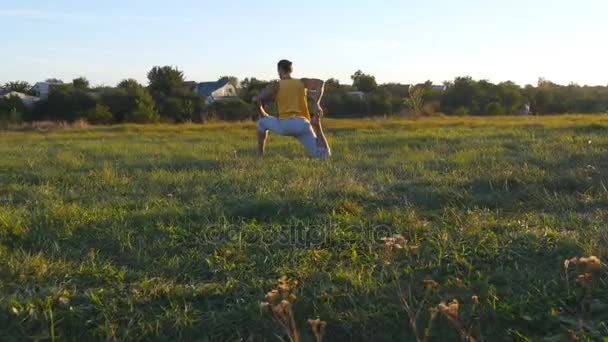 Young man sitting at green grass in the meadow and doing yoga exercise. Muscular guy stretching his body at nature. Training outdoor at sunny day. Beautiful landscape at background. Close up — Stock Video