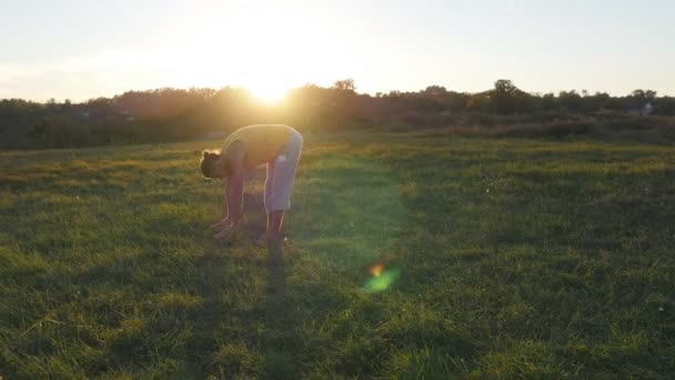Молодой человек практикует йогу движется и позиции на зеленой траве на лугу. Спортивный парень стоит в позе йоги на природе. Пейзаж как фон. Здоровый активный образ жизни йоги на свежем воздухе — стоковое видео