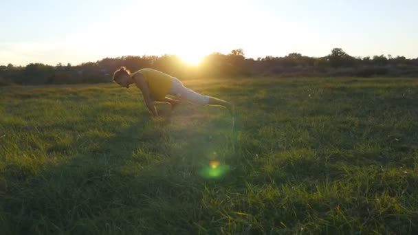 Mladý muž cvičení jógy se pohybuje a přesunete na zelené trávy na louce. Sportovní chlap stojící na jóga představuje v přírodě. Krásná krajina jako pozadí. Zdravý aktivní životní styl jogína venkovní — Stock video