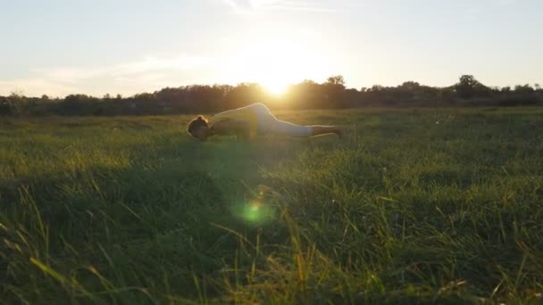 Jovem praticando movimentos de ioga e posições na grama verde no prado. Tipo desportivo em pé na pose de ioga na natureza. Bela paisagem como fundo. Estilo de vida ativo saudável de yogi ao ar livre — Vídeo de Stock