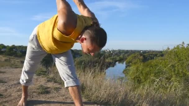 Sportieve jongeman permanent op yoga pose buiten. Kaukasische guy beoefenen van yoga beweegt en posities in de natuur. Atleet balanceren op één been. Mooi landschap als achtergrond. Gezonde actieve levensstijl — Stockvideo