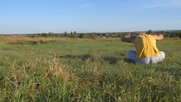 若者は草原とヨガを行使して緑の芝生に座って。筋肉男は自然に自分の体をストレッチします。晴れた日の屋外のトレーニング。バック グラウンドで美しい風景。クローズ アップ — ストック動画