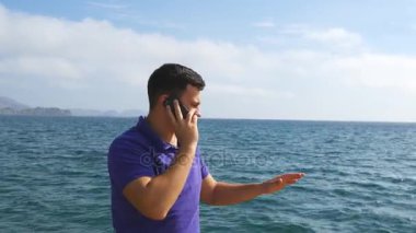 Genç yakışıklı adam üstünde hareket eden telefon bir deniz plajda konuşuyor. Ciddi adam arka plan okyanusun telefonda konuşan. Yaz tatili ara. Güzel manzara. Yakın çekim