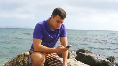 Genç adam bir taş üzerinde oturan ve denize yakın smartphone kullanarak. Yakışıklı olan ile hareket eden telefon okyanus plaj portre. Çocuk telefon app arıyorum. O mesaj sms ya da sosyal ağ sohbet