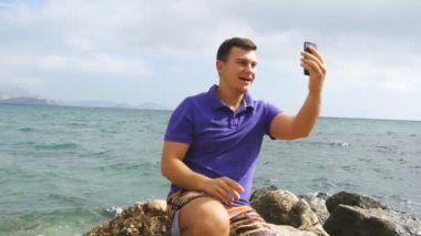 Video aramasý deniz plajda akıllı telefon olan genç gülümseyen adam. Mutlu adam video çevrimiçi sohbetler güzel manzara arka plan yapma. Çocuk cep ve gösteren okyanus üzerinde konuşuyor. Selfie yapan erkek