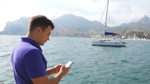 Ο νεαρός στέκεται κοντά στη θάλασσα και χρησιμοποιώντας το smartphone. Σκάφος στη θάλασσα, σε φόντο. Πορτραίτο του όμορφος άντρας με το κινητό στο ocean beach. Αυτός γραπτών μηνυμάτων sms ή να συνομιλεί στο κοινωνικό δίκτυο. Κοντινό πλάνο — Αρχείο Βίντεο