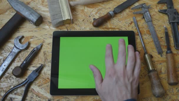 Soporte de tableta PC en modo paisaje con mesa de taller con herramientas de carpintería en el fondo. Mano de hombre usando tableta con pantalla verde. Vista superior — Vídeo de stock