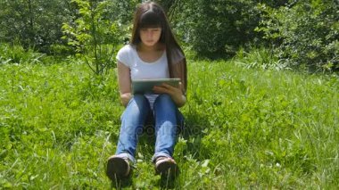 Çayır parkta çimlere oturup ve dijital tablet pc kullanarak genç beyaz esmer kız. Mutlu kadın tablet ekranına bakarak