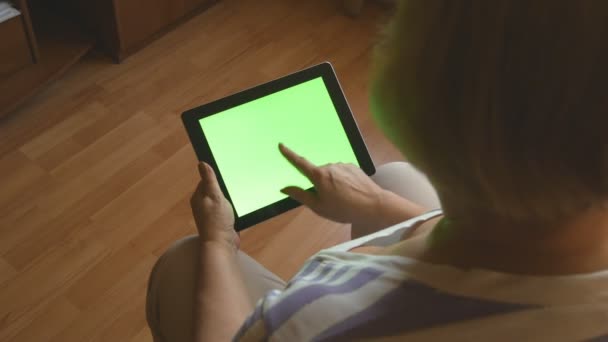 Mujer mayor sentada en el sofá en casa y utilizando una tableta digital con pantalla verde, vista trasera. Tablet PC en manos de una mujer adulta — Vídeos de Stock