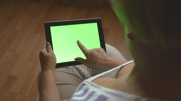 Idős asszony ül a kanapén, otthon, és a zöld képernyő, digitális tábla-pc segítségével hátulnézet. Tábla-Pc egy felnőtt nő kezet — Stock videók