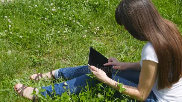 Молодая кавказская брюнетка сидит на траве на лугу в парке и пользуется цифровым планшетным ПК. Счастливая женщина смотрит на экран планшета — стоковое видео
