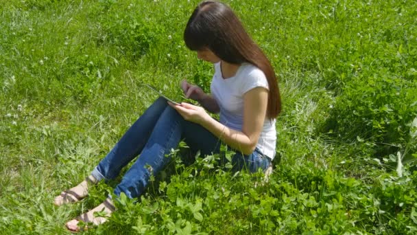 Joven chica morena caucásica sentado en la hierba en el prado en el parque y el uso de tableta digital PC. Mujer feliz mirando la pantalla de la tableta — Vídeo de stock