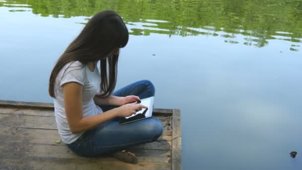 Дівчина використовує планшетний ПК і відпочиває біля озера, сидячи на краю дерев'яної дрібнички біля водної поверхні — стокове відео