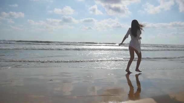 Щаслива жінка біжить і крутиться на пляжі біля океану. Молода красива дівчина насолоджується життям і розважається на морському березі. Літні канікули або відпустка. Природний ландшафт на фоні. Повільний рух — стокове відео
