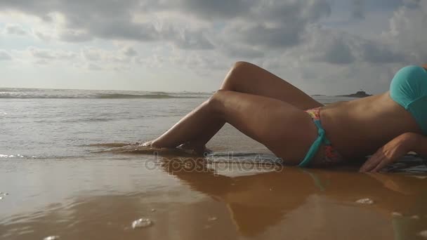 Молодая сексуальная женщина лежит на золотистом песке на морском пляже и отдыхает во время летних отпусков. Океанские волны омывают загорелые женские ноги. Медленное движение — стоковое видео