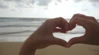 Kız güzel altın gün batımı ile deniz arka plan üzerinde kalp elleri yapma. Kadın kol içinde gündoğumu ile kalp şeklinde silüeti. Tatil kavramı. Yaz tatillerini plaj üzerinde. Ağır çekim