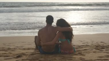 Genç romantik çift güzel günbatımı sahilde oturan ve sarılma keyif alıyor. Bir kadın ve bir adam oturuyor birlikte deniz kıyısında kum okyanus ve manzara hayran. Slowmotion yakından