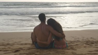 Genç Romantik Çift sahilde oturan ve sarılma harika bir manzara keyif alıyor. Bir kadın ve bir adam oturuyor birlikte sahil üzerinde kum okyanus ve manzara hayran. Ağır çekim yakın Up