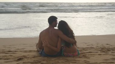 Genç Romantik Çift sahilde oturan ve akşam saatlerinde sarılma harika bir manzara keyif alıyor. Bir kadın ve bir adam oturuyor birlikte deniz kıyısında kum okyanus ve manzara hayran. Slowmotion
