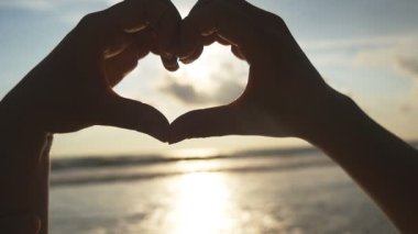 Kız güzel altın gün batımı ile deniz arka plan üzerinde kalp elleri yapma. Kadın kol içinde gündoğumu ile kalp şeklinde silüeti. Tatil kavramı. Yaz tatillerini plaj üzerinde. Ağır çekim