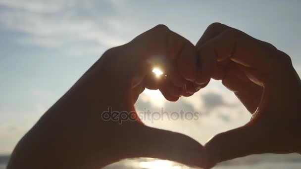 Chica haciendo corazón con sus manos sobre el fondo del mar con hermosa puesta de sol de oro. Silueta de brazo femenino en forma de corazón con salida del sol en el interior. Concepto de vacaciones. Vacaciones de verano en playa. Movimiento lento — Vídeos de Stock