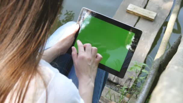 湖のほとりに木製の桟橋の上に座って緑色の画面でデジタル タブレット pc を使用して女性の手. — ストック動画