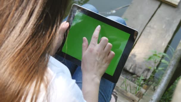 Female hands using digital tablet pc with green screen sitting on a wooden jetty by the lake. — Stock Video