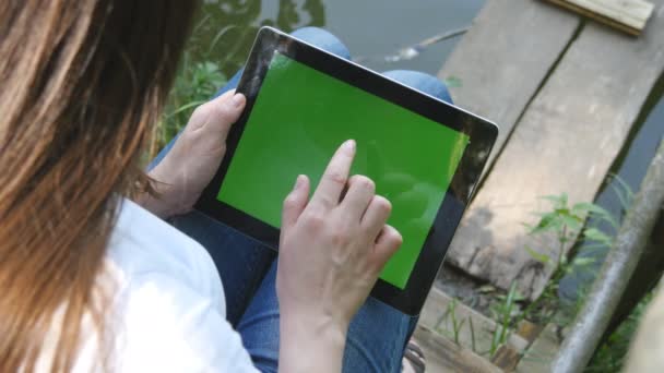 Female hands using digital tablet pc with green screen sitting on a wooden jetty by the lake. — Stock Video