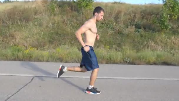 Mladý silný muž běžící na silnici v krásné přírodě. Mužské běžec běhání během cvičení školení na venkovské silnici při západu slunce. Hezký sportovní pes cvičit v létě. Trénink venku. Životní styl — Stock video