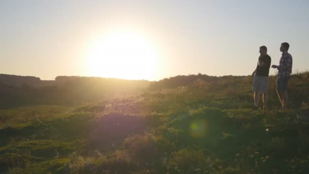 Porträtt av far med sin son prata utomhus i solnedgången. Två vuxna män står på grönt gräs på kullen och tala med varandra. Far och son spendera tid tillsammans på naturen i kväll — Stockvideo