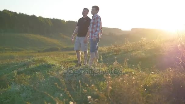 Porträt eines Vaters mit seinem Sohn, der sich bei Sonnenuntergang im Freien unterhält. Zwei erwachsene Männer stehen auf grünem Gras auf dem Hügel und sprechen miteinander. Vater und Sohn verbringen abends gemeinsam Zeit in der Natur — Stockvideo