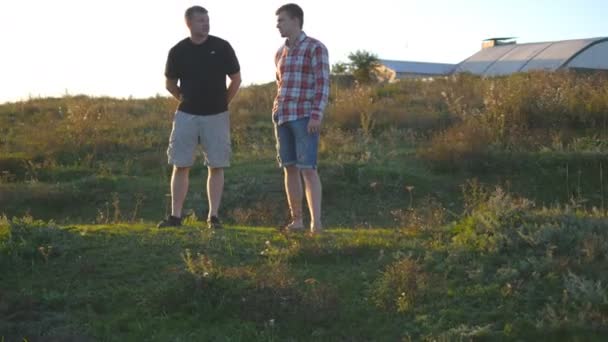 Porträtt av far med sin son prata utomhus i solnedgången. Två vuxna män står på grönt gräs på kullen och tala med varandra. Far och son spendera tid tillsammans på naturen i kväll — Stockvideo