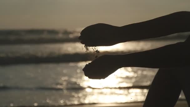 Arm van jonge vrouw spelen met zand. Vrouwelijke hand gieten zee zand door haar vingers bij zonsondergang tegen een achtergrond van de Oceaan. Slow motion close-up — Stockvideo