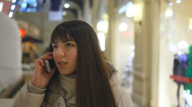 Çekici gülümseyen genç kadın kış giysileri içinde yürümek, alışveriş merkezi ve üstünde hareket eden telefon konuşma. Kız mağaza gidiş ve telefonda konuşuyor. Şirin esmer kadın alışveriş yakın çekim