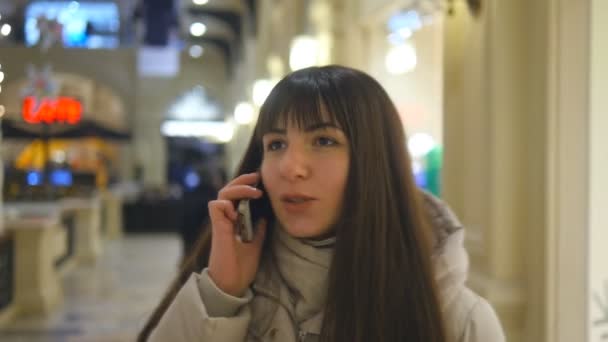 Ελκυστική χαμογελώντας νεαρή γυναίκα χειμώνα ρούχα με τα πόδια στο εμπορικό κέντρο και μιλώντας σε κινητό τηλέφωνο. Κορίτσι πηγαίνει μαζί στο κατάστημα και να μιλάει στο κινητό. Κοντινό πλάνο γλυκουλα Μελαχροινή θηλυκό αγοραστής — Αρχείο Βίντεο