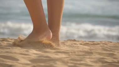 Sahilde altın kumda yürürken arka planda okyanus dalgaları olan kadın ayaklarına yakın dur. Genç kadın bacakları kuma basıyor. Deniz kıyısındaki çıplak ayaklı kız. Yaz tatili. Yavaş çekim