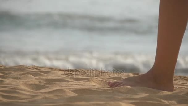 Primer plano de pies femeninos caminando sobre arena dorada en la playa con olas oceánicas al fondo. Piernas de mujer joven pisando la arena. Chica descalza en la orilla del mar. Vacaciones de verano. Movimiento lento — Vídeo de stock