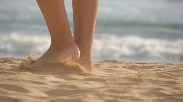 Primer plano de pies femeninos caminando sobre arena dorada en la playa con olas oceánicas al fondo. Piernas de mujer joven pisando la arena. Chica descalza en la orilla del mar. Vacaciones de verano. Movimiento lento — Vídeo de stock