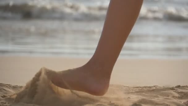 Zbliżenie żeńskich stóp spacerujących po złotym piasku na plaży z falami oceanicznymi w tle. Nogi młodej kobiety stąpającej po piasku. Boso dziewczyna na brzegu morza. Wakacje. Zwolniony ruch — Wideo stockowe