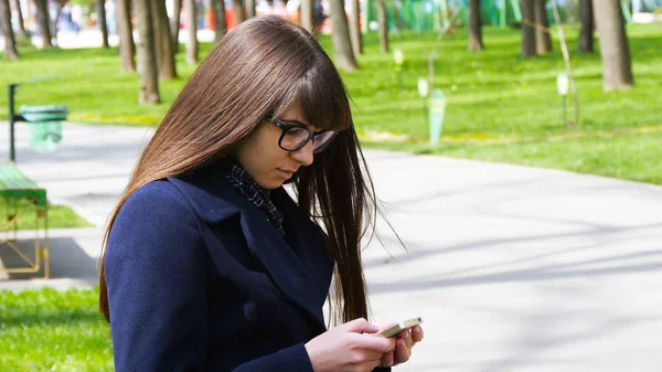 Gözlük güzel kadın hücre smartphone açık havada park - ayrıntı kullanır. Genç çekici mutlu kız bir şehir parkında rahatlatır ve bir cep telefonu kullanır. O çok mutlu ve memnun görünüyor