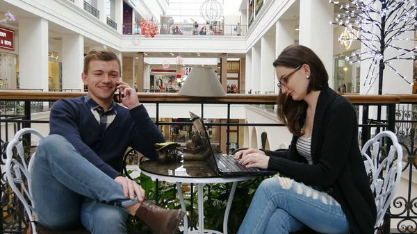 Pareja feliz con teléfono inteligente y portátil en la cafetería. Hombre hablando por teléfono, mujer trabajando en el cuaderno en la cafetería en el centro comercial . — Foto de Stock