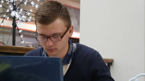 Jovem de óculos está sentado à mesa no shopping center e trabalhando em um laptop — Fotografia de Stock